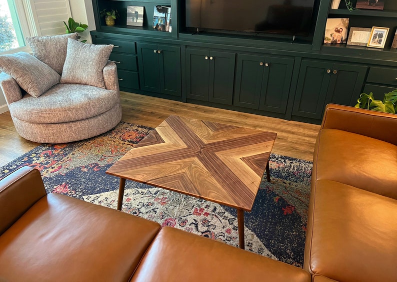 May include: A square wooden coffee table with a geometric pattern of light and dark wood. The table has a modern design and is sitting on a rug with a colorful pattern.