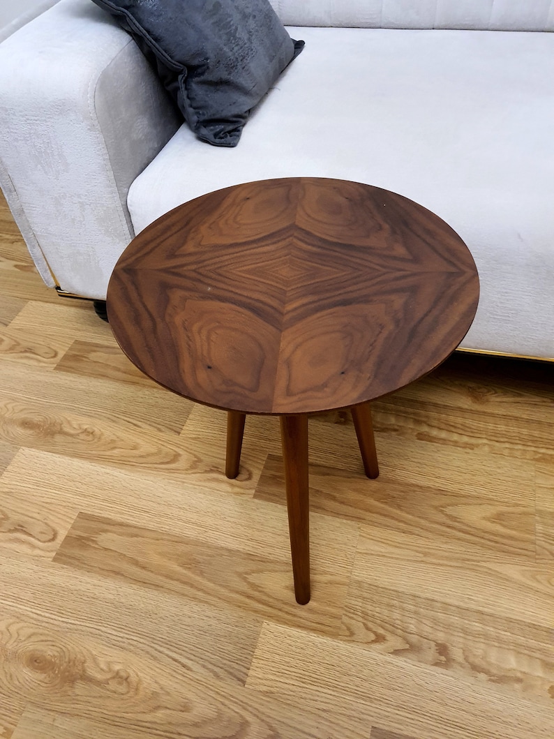 K&ouml;nnte beinhalten: Ein runder Holz-Beistelltisch mit einer dunkelbraunen Holzmaserung und drei Beinen. Der Tisch steht auf einem hellen Holzboden.