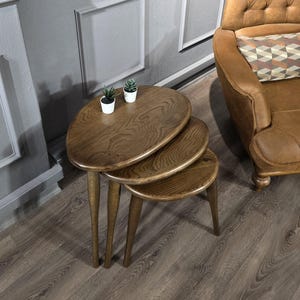 May include: Three wooden nesting tables with oval tops. The tables are brown and have a natural wood finish. The tables are arranged in a stack, with the largest table on the bottom and the smallest table on top. The tables are sitting on a wooden floor.