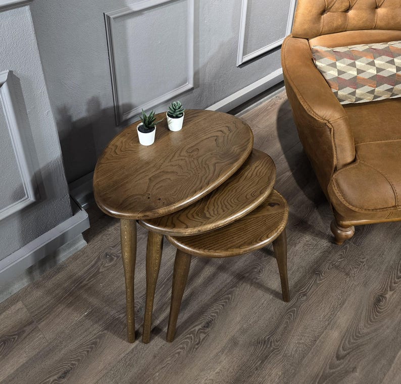 May include: Three wooden nesting tables with oval tops. The tables are brown and have a natural wood finish. The tables are arranged in a stack, with the largest table on the bottom and the smallest table on top. The tables are sitting on a wooden floor.