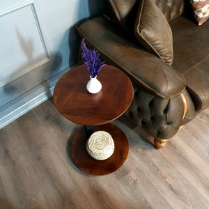 May include: A two-tiered wooden side table with a round top and a lower shelf. The table is made of dark wood and has a natural wood finish. A small white vase with purple flowers sits on the top shelf, and a woven basket sits on the lower shelf.