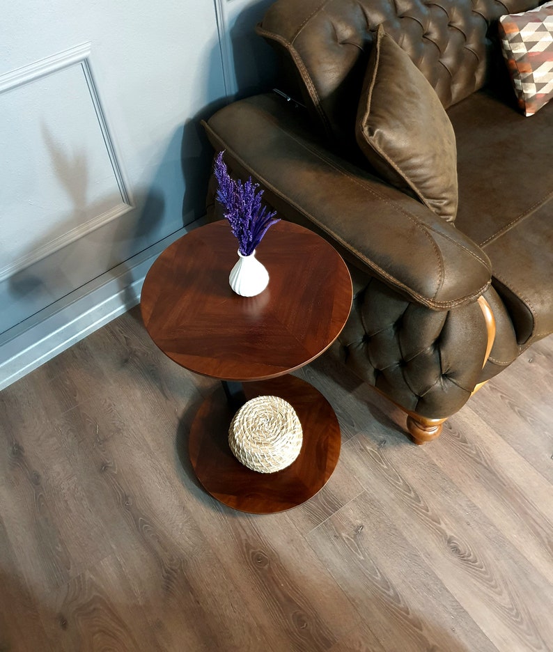 May include: A two-tiered wooden side table with a round top and a lower shelf. The table is made of dark wood and has a natural wood finish. A small white vase with purple flowers sits on the top shelf, and a woven basket sits on the lower shelf.