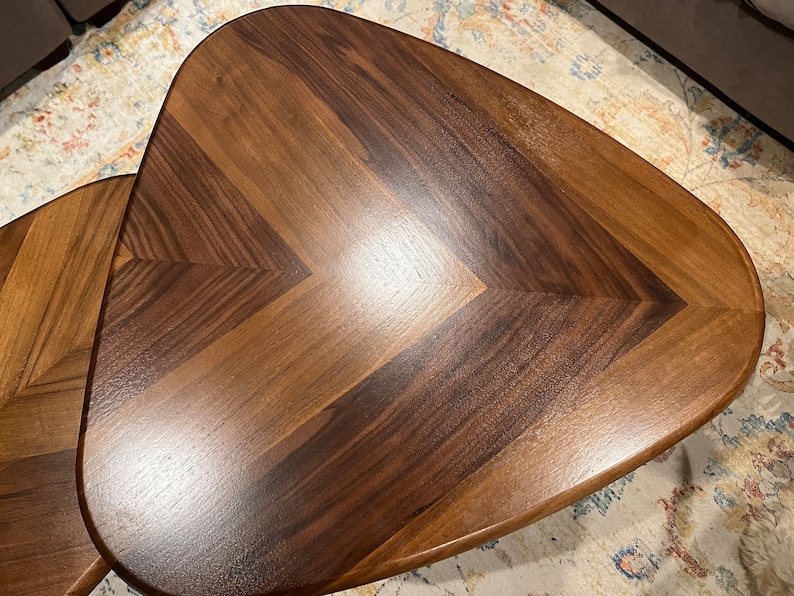 May include: Two wooden, triangular-shaped side tables with a chevron pattern. The tables are made of dark brown and light brown wood, with a glossy finish. The tables are likely used for home decor.