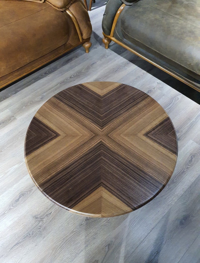 May include: A round wooden coffee table with a dark brown and light brown wood grain pattern. The table top has a geometric design with a cross shape in the center.