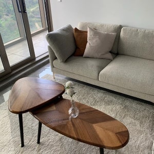 May include: Two wooden coffee tables with black legs, arranged in a layered design. The tabletops have a chevron pattern. A light gray sofa with three pillows sits behind the tables. A small glass vase with white flowers is on the lower table.