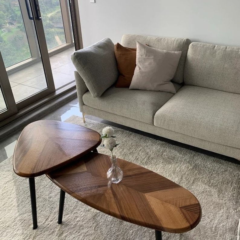 May include: Two wooden coffee tables with black legs, arranged in a layered design. The tabletops have a chevron pattern. A light gray sofa with three pillows sits behind the tables. A small glass vase with white flowers is on the lower table.