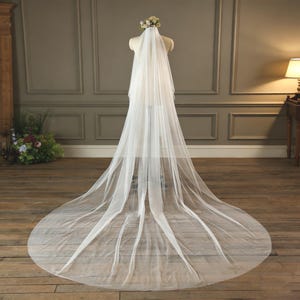 Puede incluir: Un velo de novia blanco y largo sobre un maniquí. El velo es de tela transparente y tiene un tocado floral. Se exhibe sobre un suelo de madera, con una pared de tono neutro al fondo.