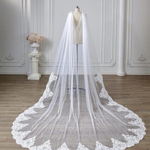 May include: A long, floor-length white bridal cape made of sheer tulle fabric. The cape features a deep V-neckline at the back and is adorned with intricate white floral lace embroidery along the entire hemline, creating a sweeping train.