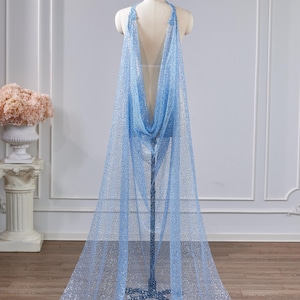 Puede incluir: Una capa azul claro y brillante con cuello halter está drapeada sobre un maniquí. La tela es transparente y llega hasta el suelo, creando un efecto fluido. La capa se exhibe en un estudio con un fondo neutro.