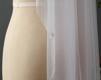 May include: A white bridal veil, sheer and flowing, with delicate beaded detailing. The veil is draped over a mannequin, highlighting its texture and design. The beaded accents add a subtle shimmer to the fabric.