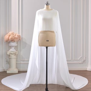 May include: A white cropped top featuring a high neckline and long, flowing sheer cape sleeves that extend to the floor, creating a dramatic train. The garment is displayed on a mannequin with the number 88 printed on the front torso section.