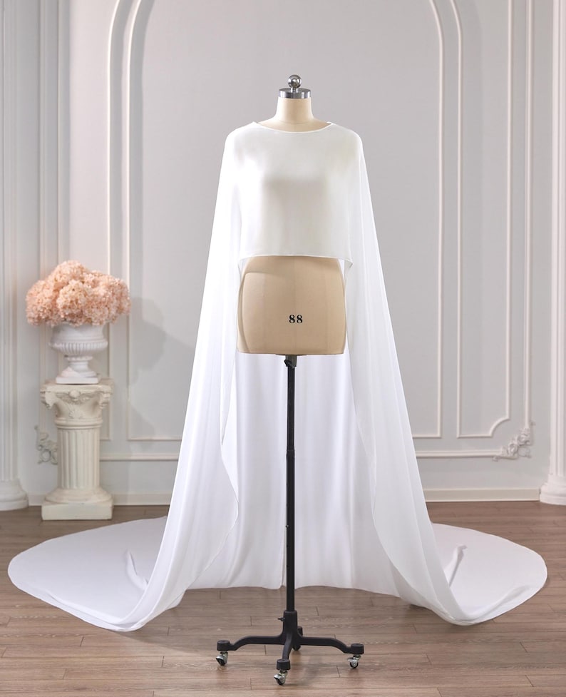 May include: A white cropped top featuring a high neckline and long, flowing sheer cape sleeves that extend to the floor, creating a dramatic train. The garment is displayed on a mannequin with the number 88 printed on the front torso section.