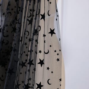 May include: A sheer black tulle fabric featuring a repeating pattern of dark black velvet stars and crescent moons. The lightweight, semi-transparent material is draped to showcase the celestial motif across the surface of the fine mesh textile.