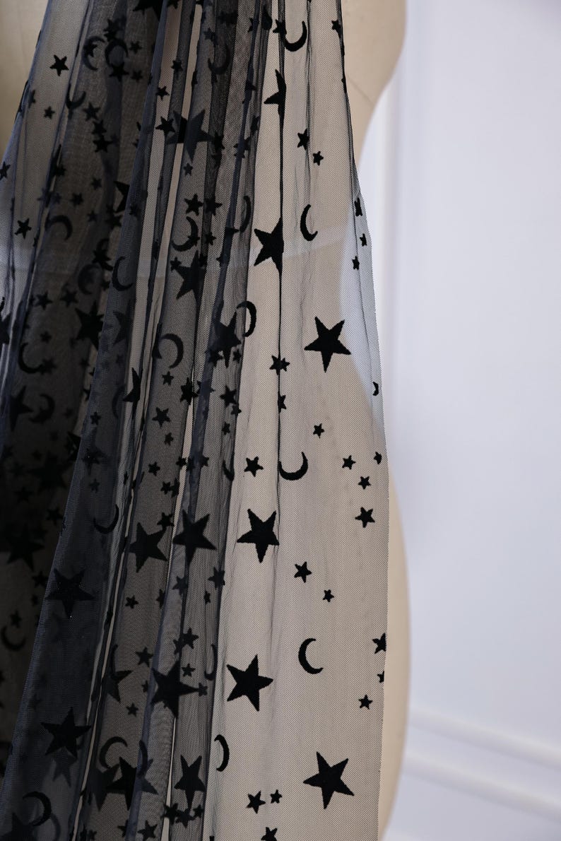 May include: A sheer black tulle fabric featuring a repeating pattern of dark black velvet stars and crescent moons. The lightweight, semi-transparent material is draped to showcase the celestial motif across the surface of the fine mesh textile.
