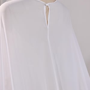 May include: A sheer white blouse featuring a round neckline and a vertical keyhole opening at the center back. The garment is secured at the top of the keyhole with a single small white button and a matching fabric loop closure.