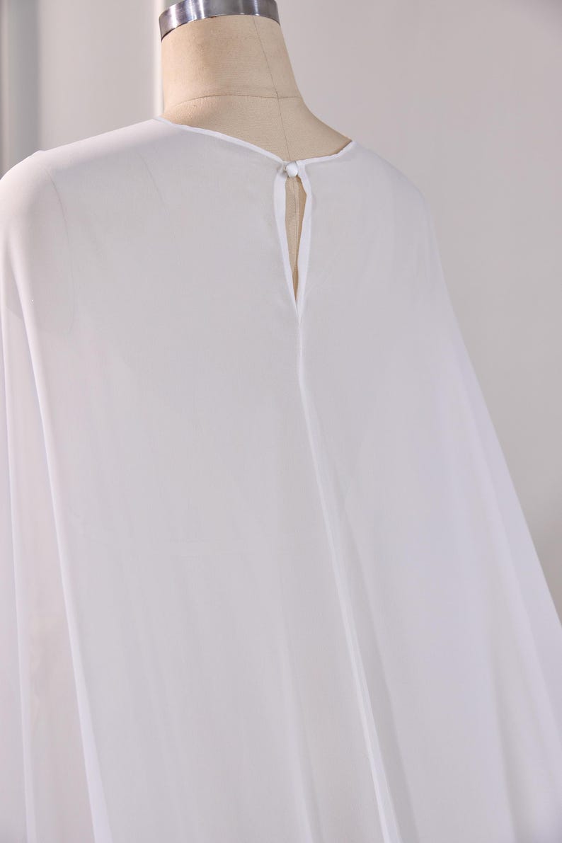 May include: A sheer white blouse featuring a round neckline and a vertical keyhole opening at the center back. The garment is secured at the top of the keyhole with a single small white button and a matching fabric loop closure.