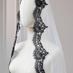 Op de afbeelding: Een witte bruidssluier met een zwarte bloemen kanten rand. De kanten details zijn zichtbaar langs de randen van de sluier, wat een delicate en elegante uitstraling creëert. De sluier wordt op een mannequin getoond.
