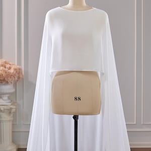 May include: A white, cropped, sleeveless cape-style top made of a sheer, flowing fabric. The garment features a high, rounded neckline and drapes down from the shoulders to create long, floor-length panels on both sides of the mannequin.