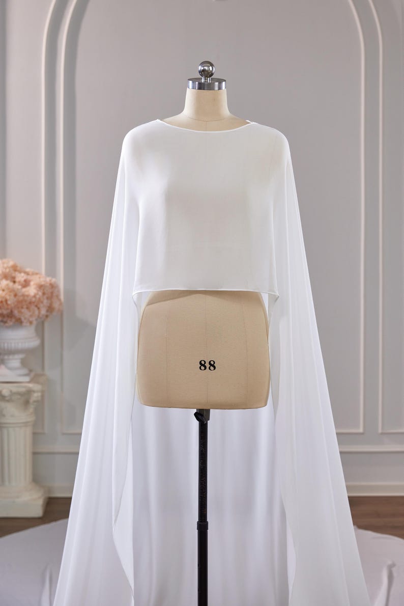 May include: A white, cropped, sleeveless cape-style top made of a sheer, flowing fabric. The garment features a high, rounded neckline and drapes down from the shoulders to create long, floor-length panels on both sides of the mannequin.