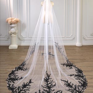 May include: A long, floor-length white tulle veil featuring intricate black floral lace embroidery along the scalloped edges and hem. The sheer fabric drapes from a central point, creating a dramatic train that spreads across the floor.
