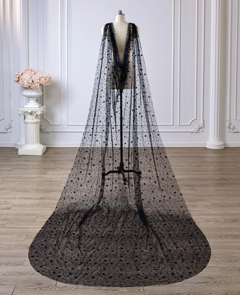 May include: A floor-length black sheer cape made of tulle fabric, featuring an all-over pattern of small black stars and crescent moons. The cape is draped over a mannequin, extending to the floor with a wide, rounded train at the base.