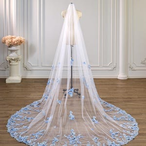 Pode incluir: Um longo véu de noiva branco que chega ao chão, feito de tecido de tule transparente. O véu é decorado com aplicações florais azul-claras espalhadas, concentradas mais densamente ao longo da borda inferior larga e arredondada.