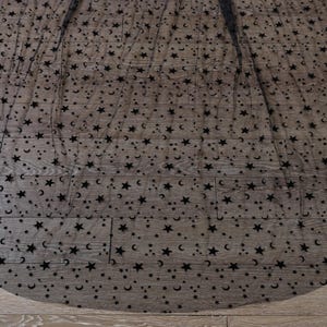 May include: A sheer black fabric overlay featuring a repeating pattern of small black velvet stars and crescent moons. The lightweight, semi-transparent material is draped in a circular shape, showcasing the dense celestial motif throughout.