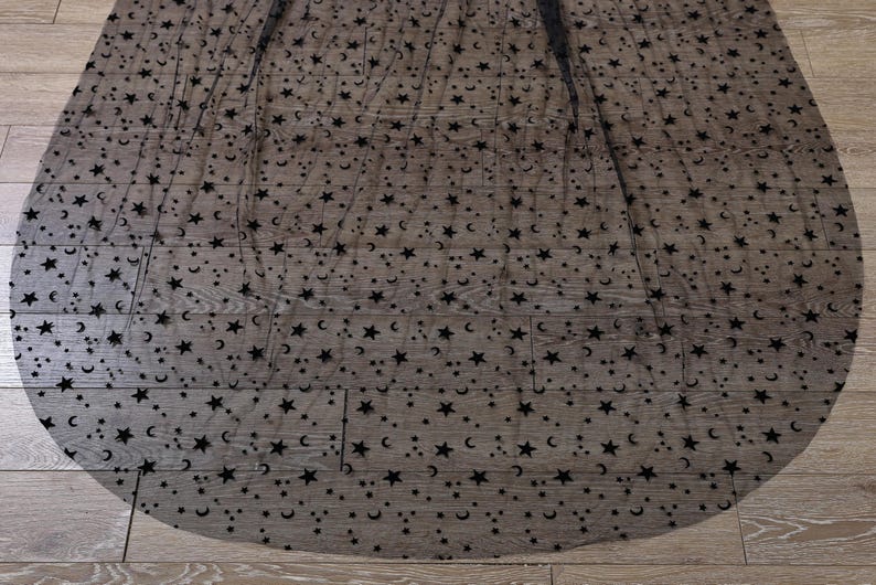 May include: A sheer black fabric overlay featuring a repeating pattern of small black velvet stars and crescent moons. The lightweight, semi-transparent material is draped in a circular shape, showcasing the dense celestial motif throughout.