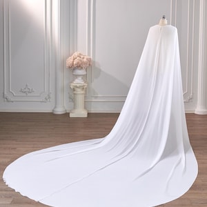 May include: A floor-length white cape made of flowing, lightweight fabric. The garment features a rounded neckline with a small button closure at the top, draping down from a mannequin to form a long, circular train that spreads across the floor.