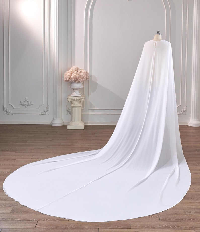 May include: A floor-length white cape made of flowing, lightweight fabric. The garment features a rounded neckline with a small button closure at the top, draping down from a mannequin to form a long, circular train that spreads across the floor.