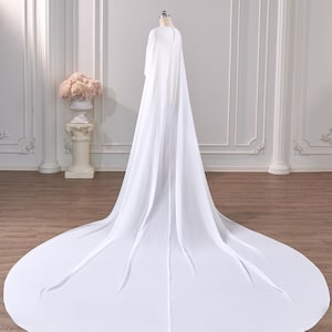 May include: A floor-length white cape made of flowing, lightweight fabric. The garment features a high neckline with a small button closure at the back and a long, sweeping train that extends across the floor in a wide, circular shape.
