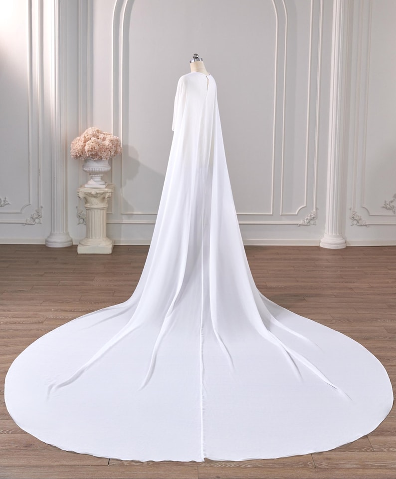 May include: A floor-length white cape made of flowing, lightweight fabric. The garment features a high neckline with a small button closure at the back and a long, sweeping train that extends across the floor in a wide, circular shape.