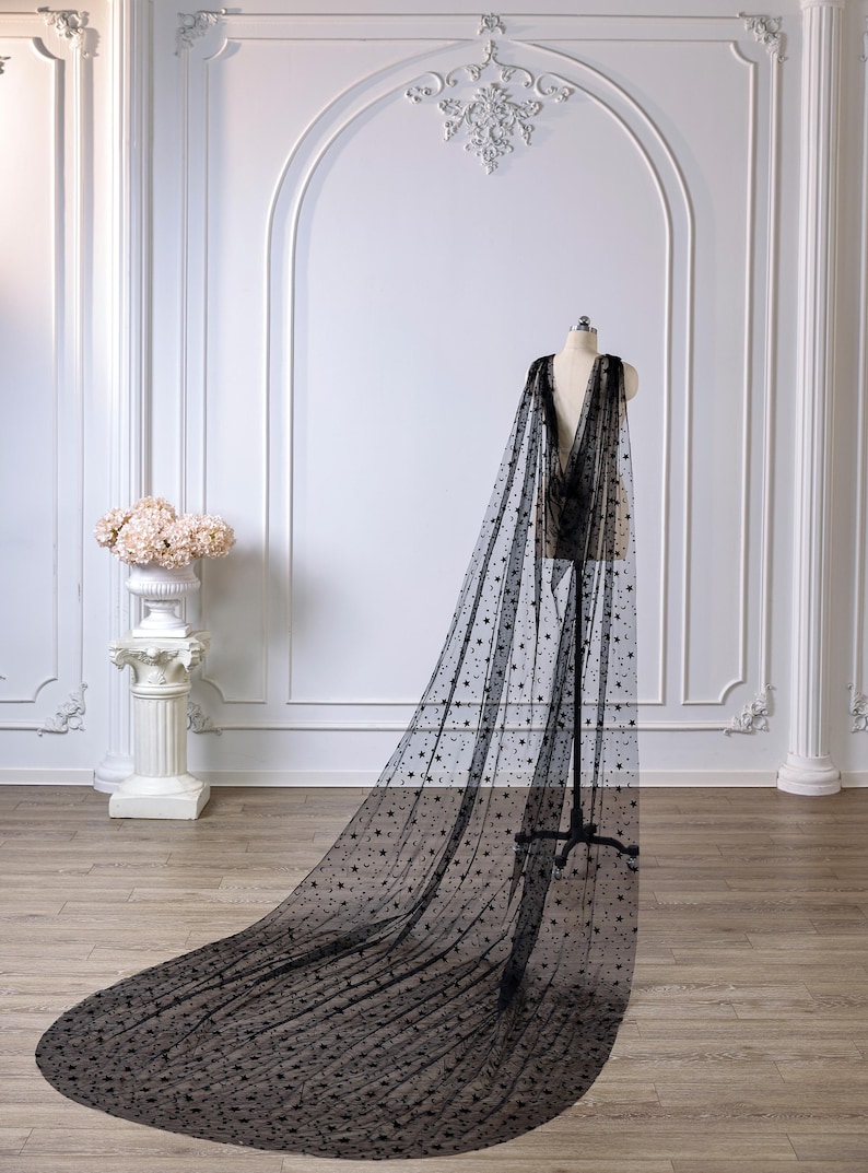 May include: A floor-length black sheer cape made of tulle fabric, featuring an all-over pattern of small embroidered stars and crescent moons. The cape is draped over a mannequin, extending onto the floor to create a long, flowing train.