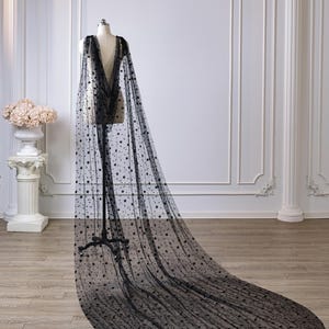 May include: A floor-length black sheer cape made of tulle fabric featuring a scattered pattern of small black stars and crescent moons. The cape drapes from the shoulders of a mannequin, extending into a long, wide train that pools on the floor.