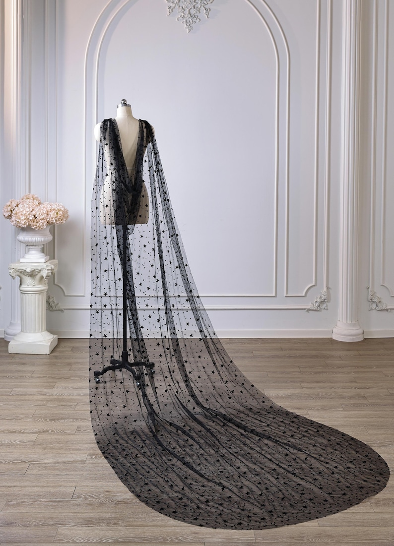 May include: A floor-length black sheer cape made of tulle fabric featuring a scattered pattern of small black stars and crescent moons. The cape drapes from the shoulders of a mannequin, extending into a long, wide train that pools on the floor.