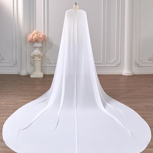 May include: A floor-length white bridal cape made of sheer, flowing fabric. The cape features a rounded neckline with a single central closure at the top, draping down the back to form a wide, circular train that spreads across the floor.