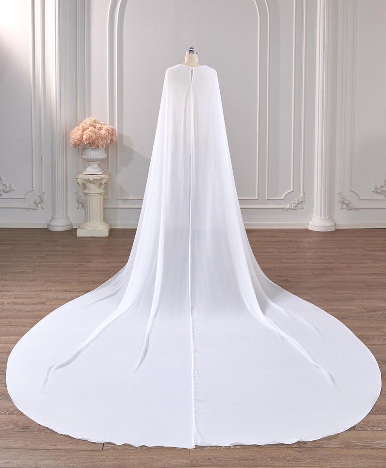 May include: A floor-length white bridal cape made of sheer, flowing fabric. The cape features a rounded neckline with a single central closure at the top, draping down the back to form a wide, circular train that spreads across the floor.