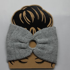May include: Gray knitted hair scrunchie with a hole in the center, displayed on a brown cardboard cutout of a woman's head.
