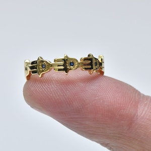Puede incluir: Anillo de oro con un patrón repetido de manos con un diseño de ojo malvado. Cada ojo está engastado con una pequeña piedra azul.