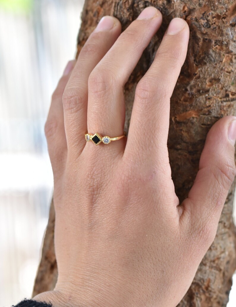 May include: Gold ring with three square-shaped stones. The center stone is black, and the stones on either side are clear.