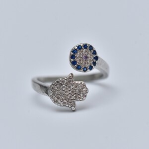 Può includere: Un anello d'argento con un motivo a mano di Hamsa e un ciondolo con l'occhio turco. Entrambi i ciondoli sono decorati con pietre di zircone cubico trasparenti e blu.