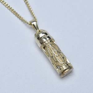 Puede incluir: Un collar de cadena de oro con un colgante con forma de cilindro pequeño. El colgante tiene un diseño intrincado de una mano con un patrón detallado.