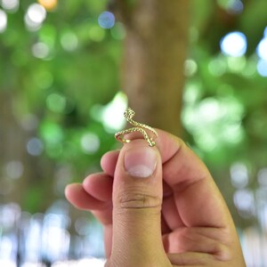 May include: A gold snake ring with a textured design. The ring is held by a hand against a blurred green background.