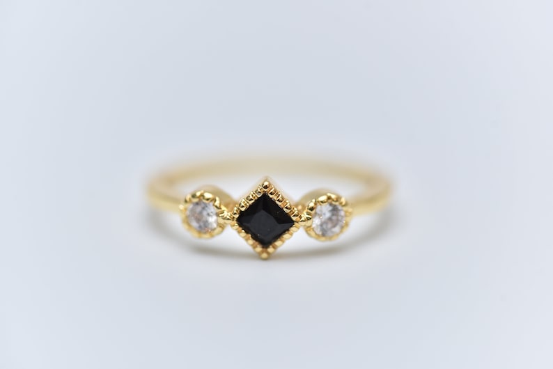 May include: A gold ring with three stones. The center stone is a black square, and the two stones on either side are round and white.