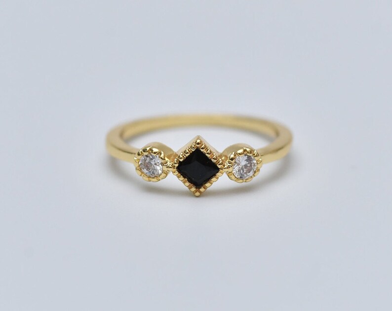 May include: A gold ring with three stones. The center stone is a black square, and the two stones on either side are round and white.