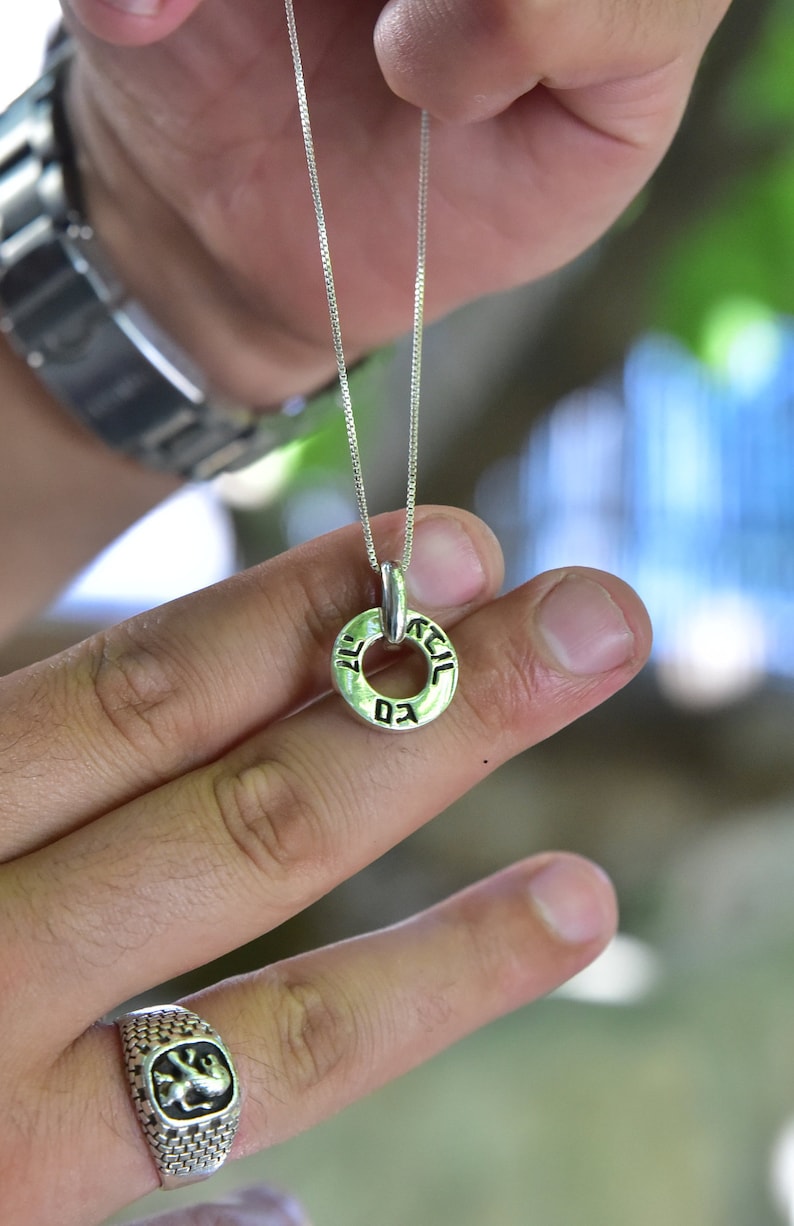 May include: A silver chain necklace with a round pendant inscribed with Hebrew text. The pendant is held by a person's hand, and a silver ring with a lion design is on a finger on the same hand.