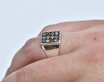May include: Silver ring with a square face featuring a grid of colorful gemstones. The gemstones include red, blue, green, yellow, and pink. The ring is on a hand.