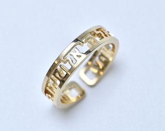 May include: A gold-colored adjustable ring with Hebrew lettering cut out along the band. The ring has a rectangular shape with rounded edges and is set against a white background. The ring is a piece of jewelry.