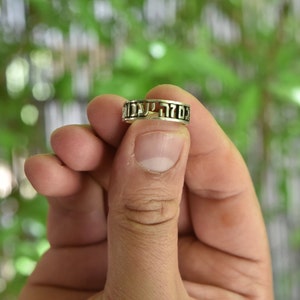 May include: A silver ring with Hebrew lettering. The ring is being held by a finger.