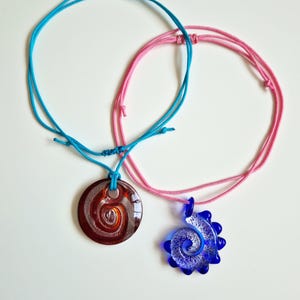 May include: Two necklaces with spiral pendants. One pendant is a dark red and brown swirl on a blue cord. The other is a blue pendant with a white swirl on a pink cord. Both cords are adjustable.